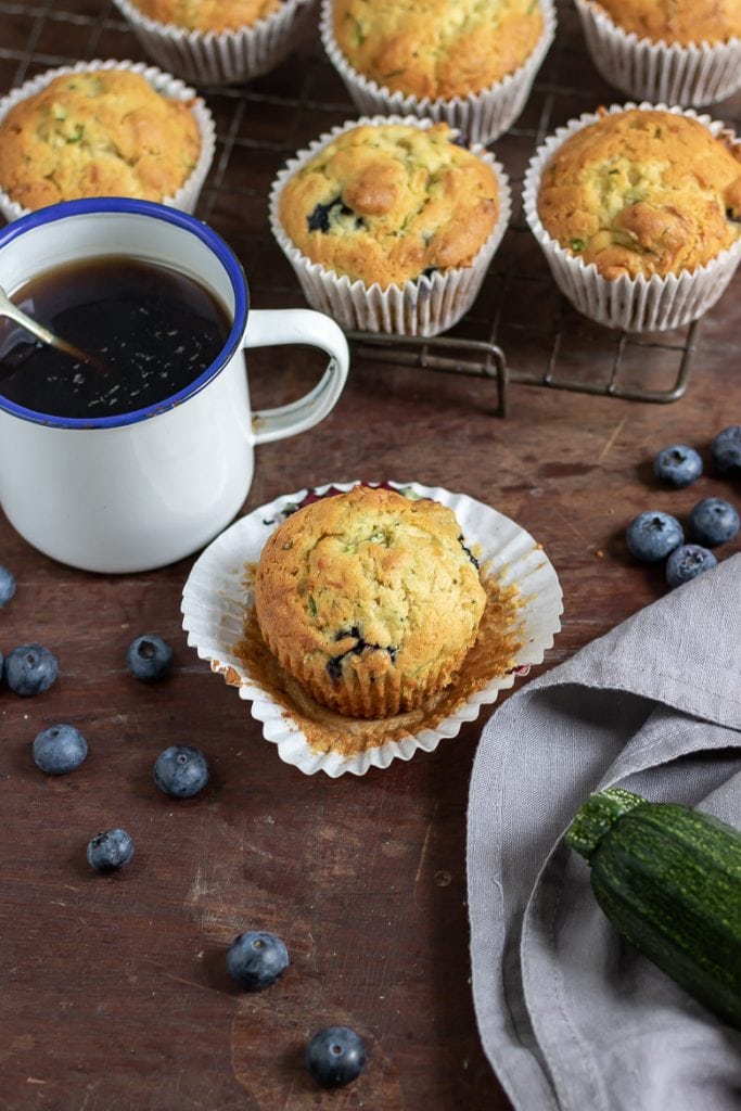 A zucchini muffin next to mug of coffee and other courgette muffins. Blueberries scattered around.