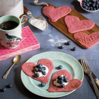 Heart-shaped beet pancakes with yogurt and blueberries on a plate surrounded my a mug, more heart pancakes and blueberries