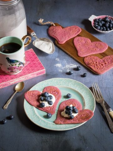 Heart-shaped beet pancakes with yogurt and blueberries on a plate surrounded my a mug, more heart pancakes and blueberries