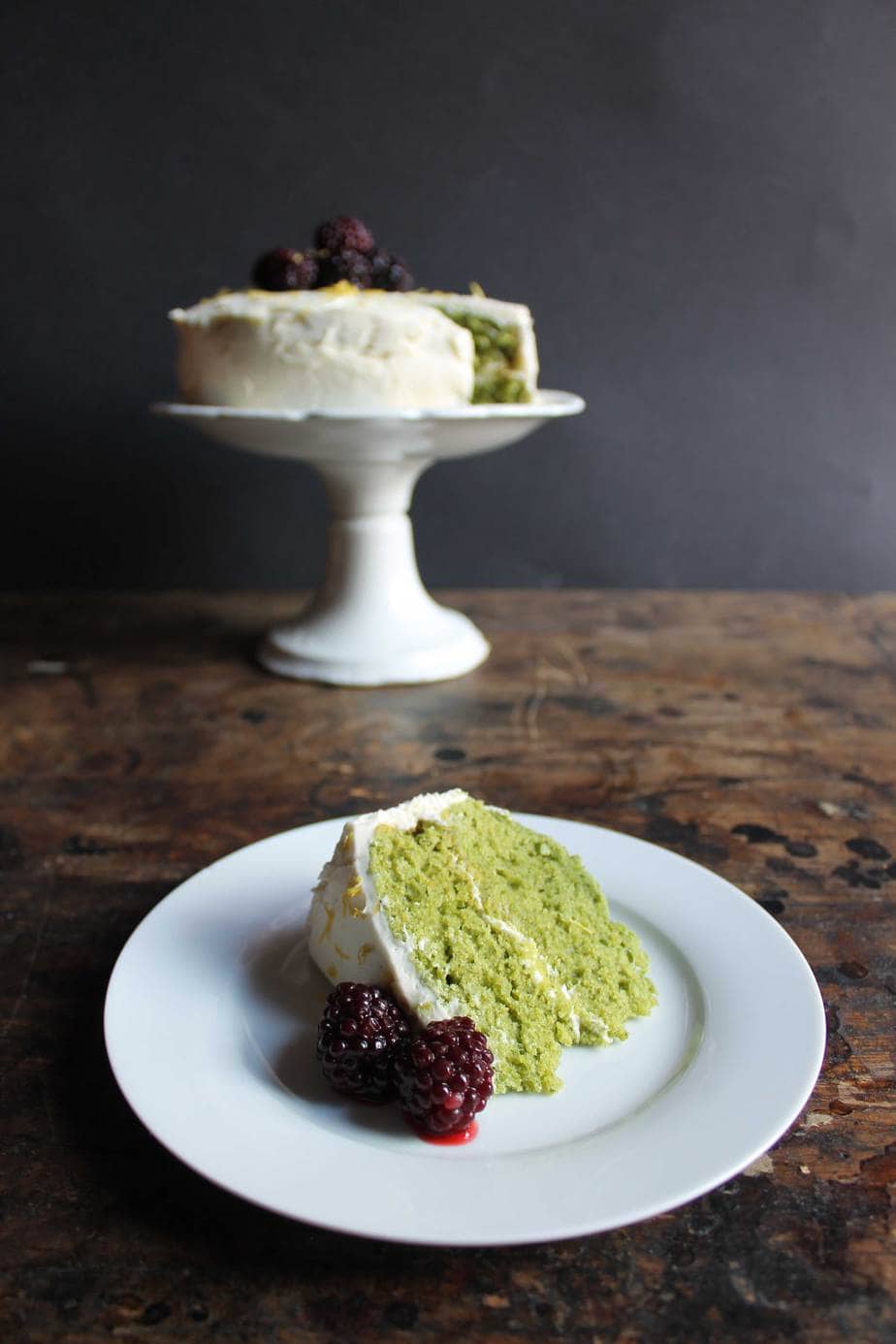 A slice of green Stinging Nettle Cake on a plate in front of the cake on a cake stand.