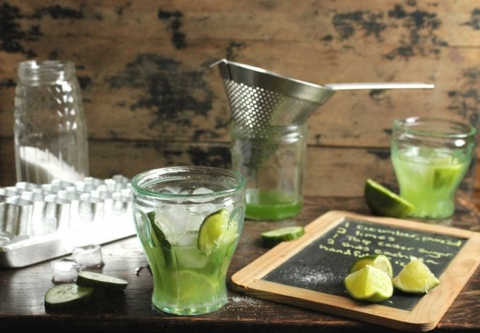 Cucumber Caipirinha Cocktail in a glass next to ice, a sieve and recipe on chalkboard