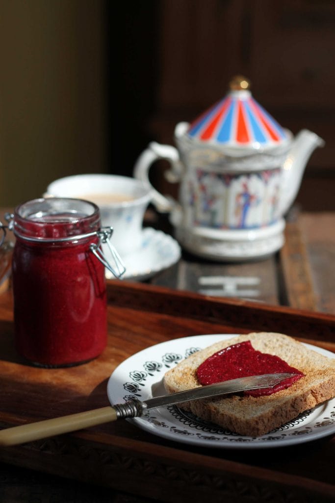 A jar of raspberry beet chia jam, plus some spread on toast, in front of a teapot and cup of tea.