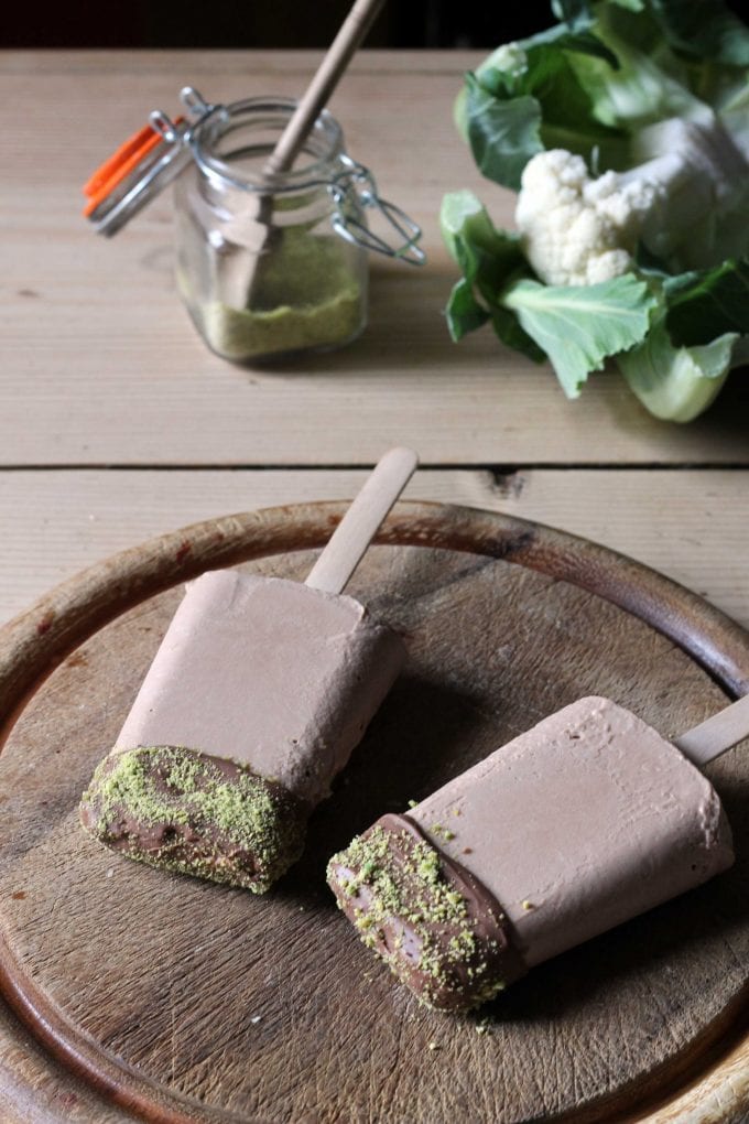 Cauliflower and Chocolate Ice Lollies with Pistachio Dust on a wooden board next to a head of cauliflower.