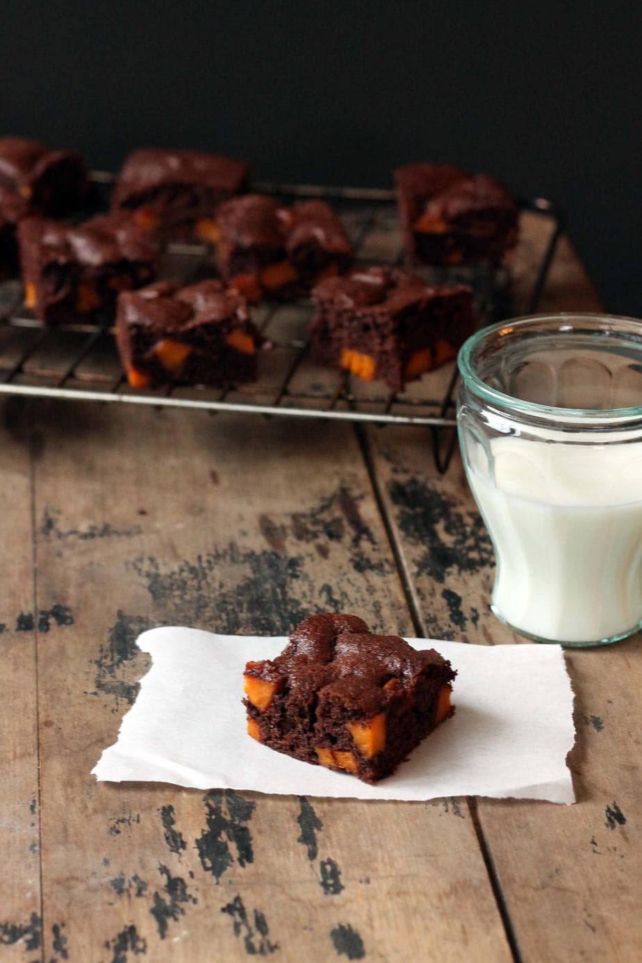 A piece of brownie on baking paper in front of a glass of milk and a cooling rack of brownies.