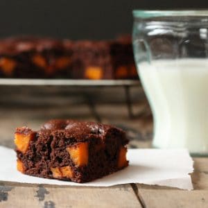 A piece of brownie on baking paper in front of a glass of milk.