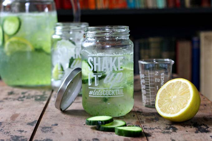 A jar glass full of fresh cucumber gin and tonic, next to slices of lime and cucumber