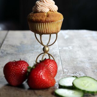 A cupcake on a golden stand with strawberries and cucumber in front of it.