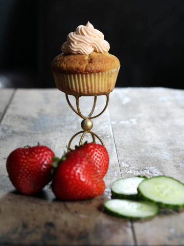 A cupcake on a golden stand with strawberries and cucumber in front of it.  Apple Parsnip Cupcakes with Boozy Apple Ci CucumberPimmsCupcakes 360x480