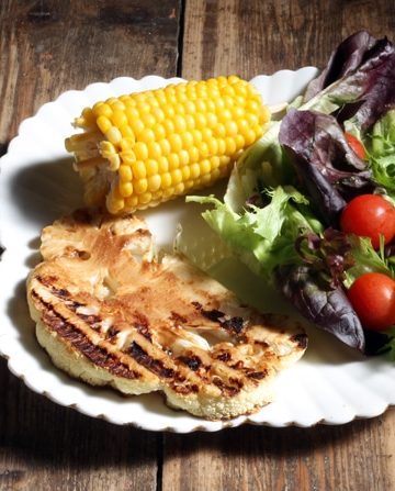 A table with a plate of cauliflower steak, salad and corn on the cob.