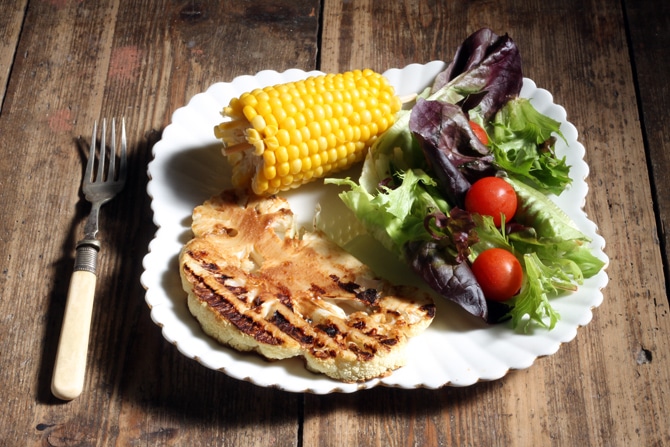 A table with a plate of cauliflower steak, salad and corn on the cob.