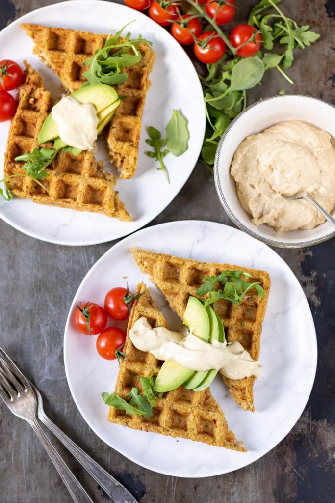 Two plates with falafel waffles, tomatoes, avocado and hummus.