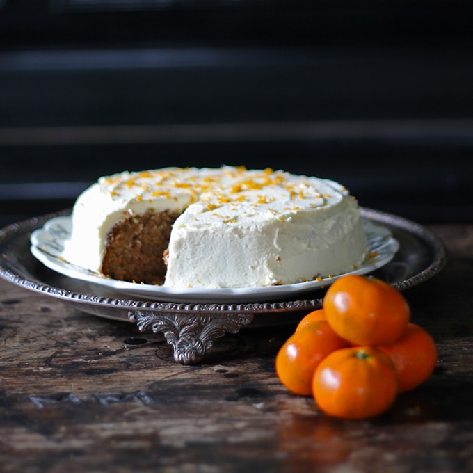 A cake with a piece taken out, on a cake stand next to a pile of tangerines.