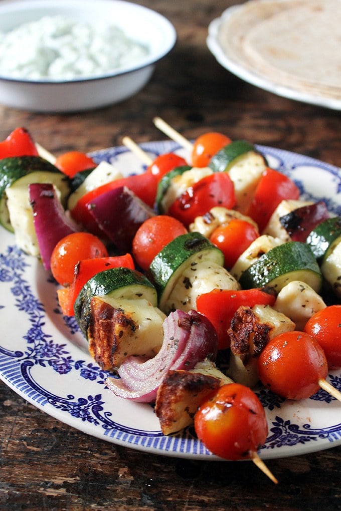 A vintage plate with skewers of grilled halloumi souvlaki with tzatziki in the background.