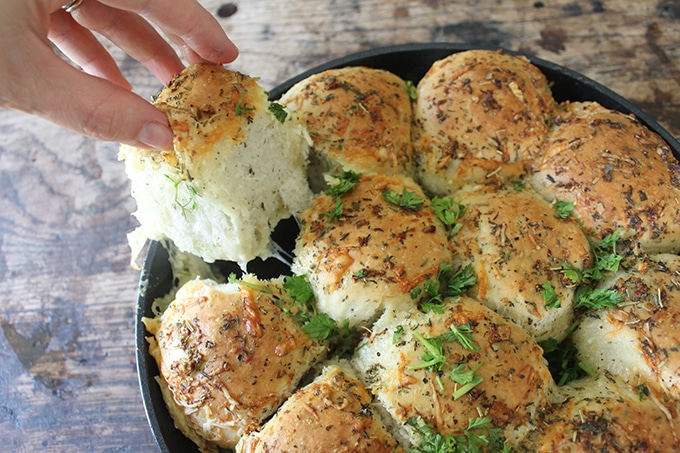 A hand taking a dinner roll from a pan of fresh Cheesy Garlic Skillet Rolls - with loads of oozy cheese pulling from it. Get the recipe now!