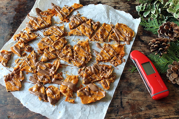 Ginger Pecan Brittle with Chocolate Drizzle. Recipe is shown on white baking paper next to food thermometer, pine cones and Christmas greens. 