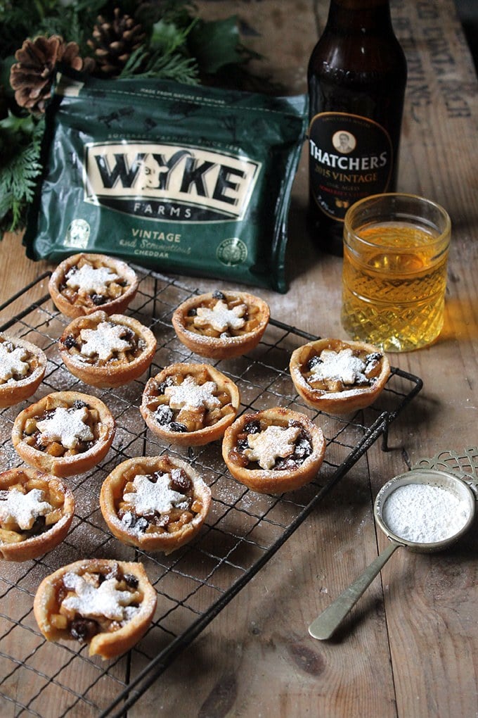 Wooden table with mince pies on a wire rack, with a block of cheese and bottle of cider.