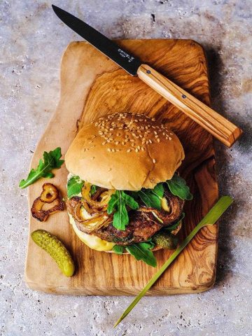 Burger on a wooden cutting board, with a pickle and knife next to it.