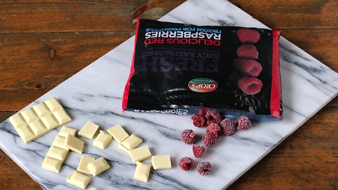 A bag of frozen raspberries next to squares of white chocolate on a marble cutting board. 