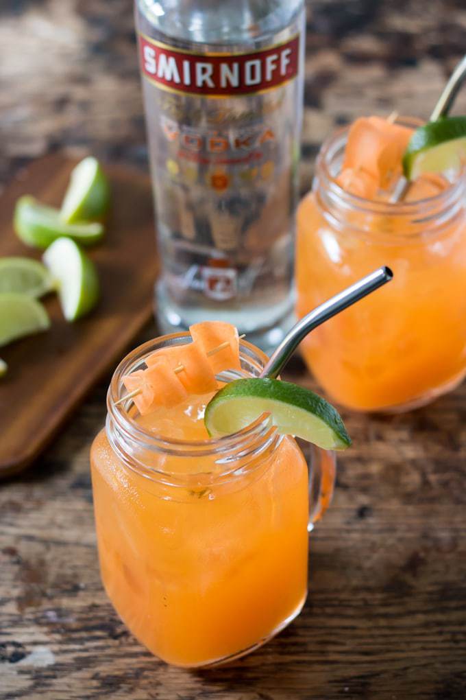 Two glasses of Carrot Moscow Mule cocktail in front of a bottle of vodka