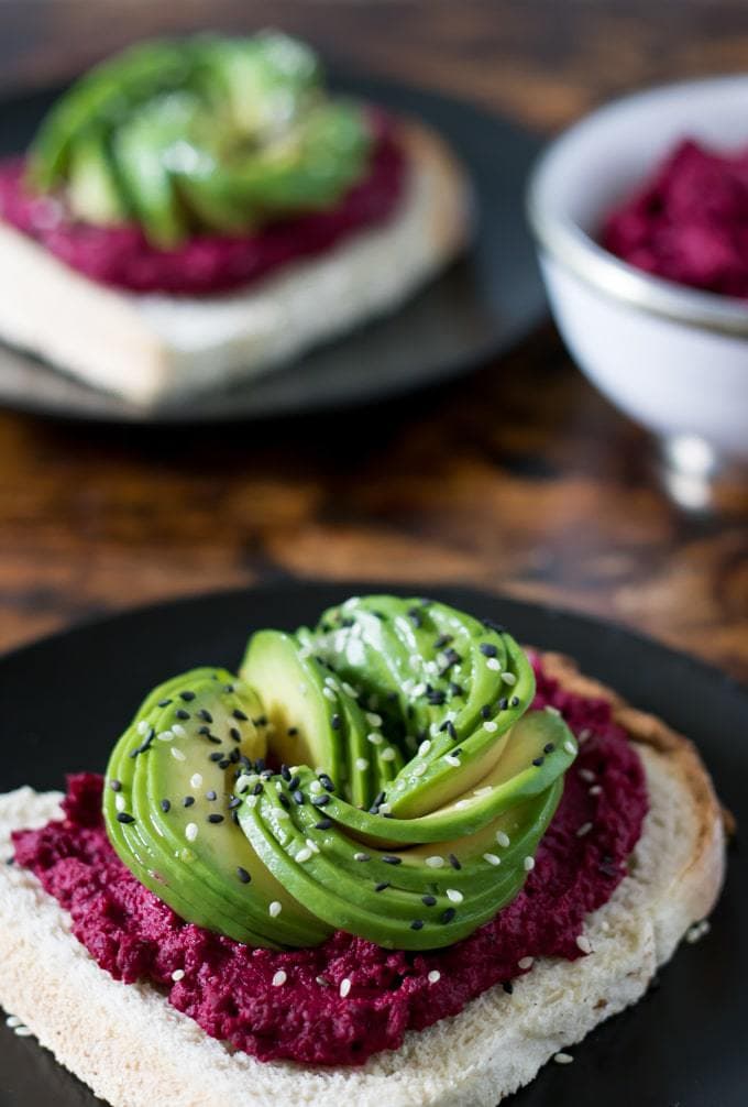 Close up of toast spread with beet hummus and topped with avocado sliced into a rose shape, then sprinkled with black and white sesame seeds. 
