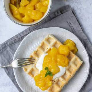 Mango compote made with fresh or frozen mango on a waffle on a white plate on a grey napkin next to a bowl of compote.
