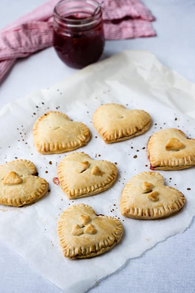 Baking paper with heart shaped jam-filled hand pies and a pot of strawberry jam. 