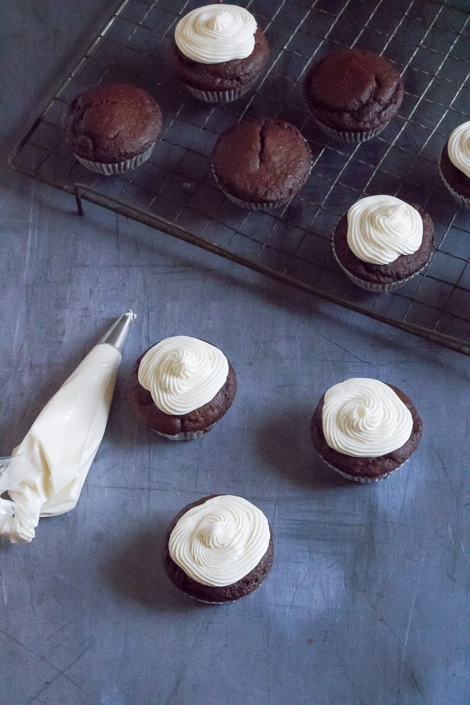 A cooling tray of easy vegan chocolate cupcakes being frosted with vanilla frosting