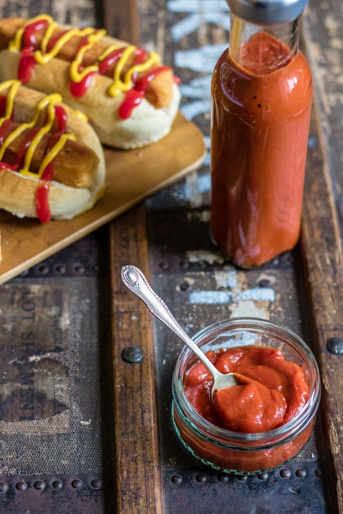 A bottle and dish of quick and easy homemade tomato ketchup with hot dogs in the background. 