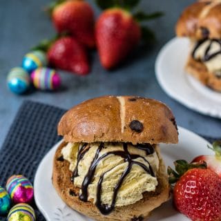 Three ingredient hot cross bun ice cream sandwiches served on a plate with a drizzle of chocolate sauce and a few strawberries.