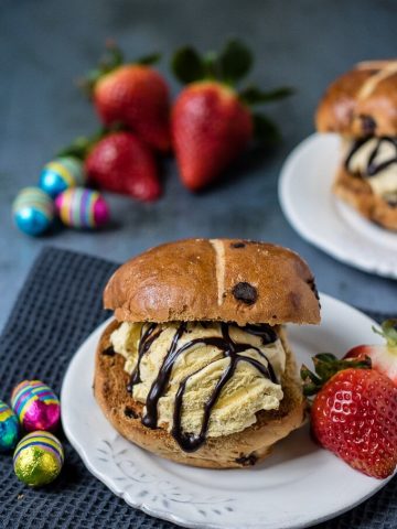 Three ingredient hot cross bun ice cream sandwiches served on a plate with a drizzle of chocolate sauce and a few strawberries.