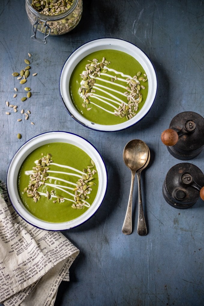 Two bowls of kale soup served with seeds and a drizzle of cream, on a table with vintage spoons and salt and pepper shakers.