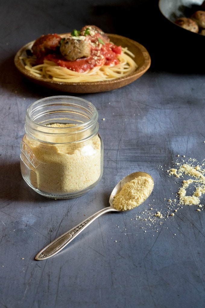 A jar of easy cheesy vegan parmesan cheese, made with cashes and spices, with a plate of spaghetti and aubergine meatballs in the background