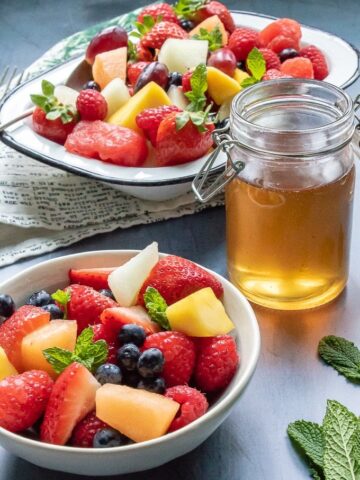 Two bowls full of fresh fruit salad and sprigs of mint, next to a jar of quick and easy tea simple syrup (sugar syrup infused with tea leaves)