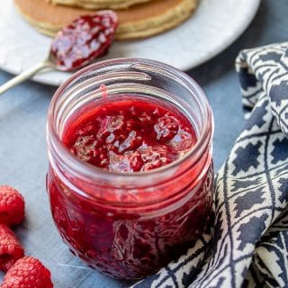 Close up of a jar of thick raspberry compote next to a stack of pancakes to spoon it onto.