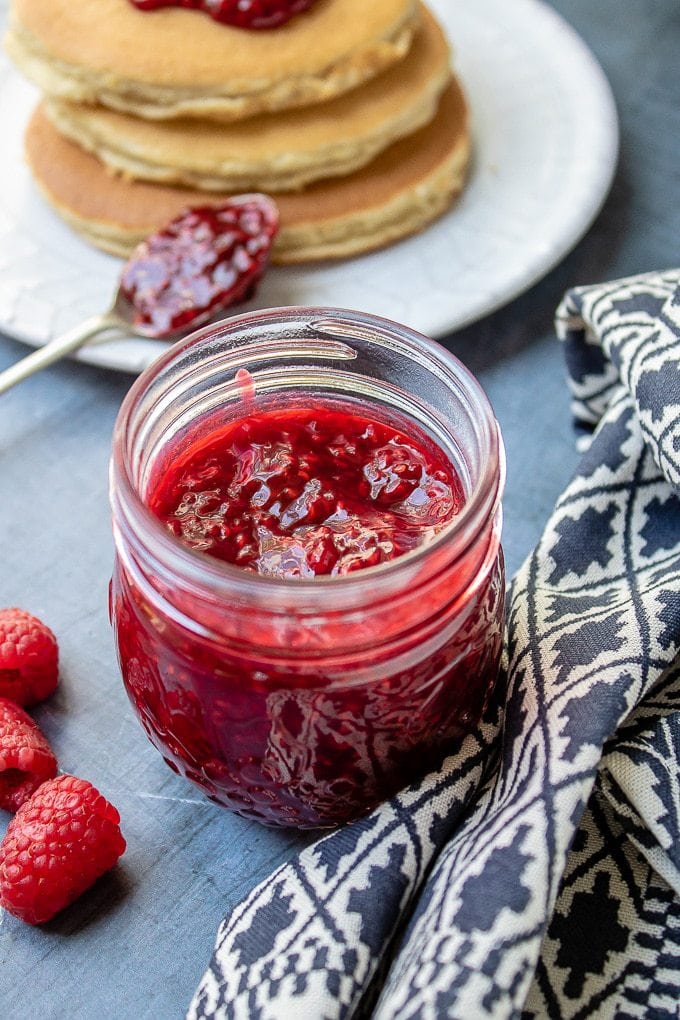 Close up of a jar of thick raspberry compote next to a stack of pancakes.