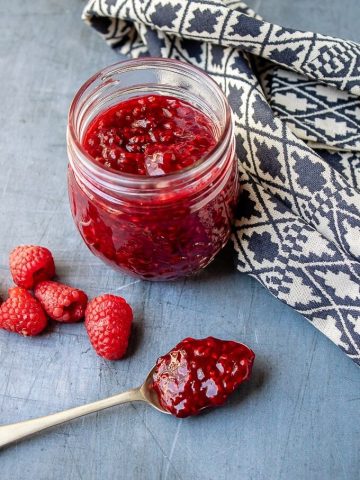 A jar of quick, easy and tasty raspberry compote, with a spoonful of it next to a blue and white tea towel.