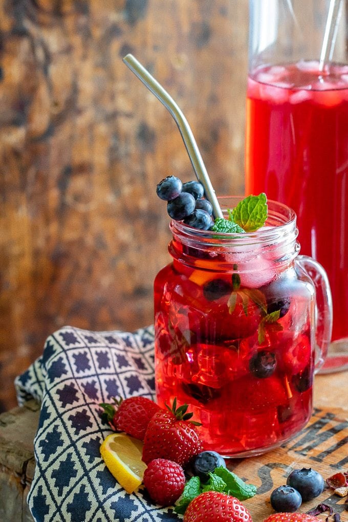 Close up of a glass of berry iced tea, with berries and mint in the glass with ice and a jug of berry iced tea in the background.