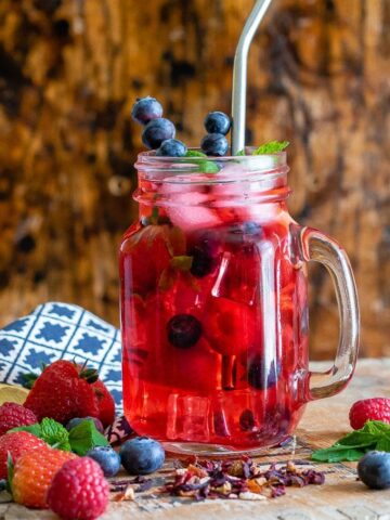 A glass of berry iced tea made with berry loose tea, ginger and berries. With berries on the table and sprigs of mint.