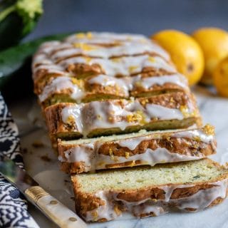 Sliced loaf of courgette cake topped with lemon drizzle. On a marble slab next to vintage knife, blue tea towel and lemons.