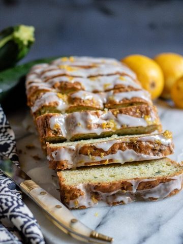 Sliced loaf of courgette cake topped with lemon drizzle. On a marble slab next to vintage knife, blue tea towel and lemons.