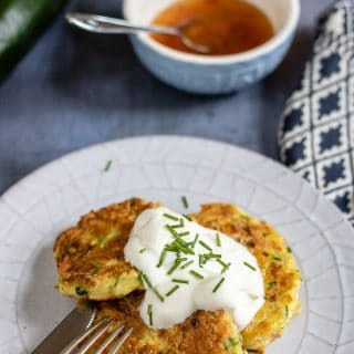 A plate with cheesy courgette fritters recipe topped with sour cream and chopped chives, next to a bowl of sweet chilli sauce.