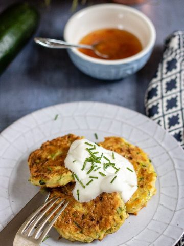 A plate with cheesy courgette fritters recipe topped with sour cream and chopped chives, next to a bowl of sweet chilli sauce.