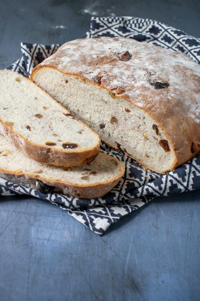 A freshly baked date and walnut bread on a blue background with patterned tea towel and two slices cut from the bread. 