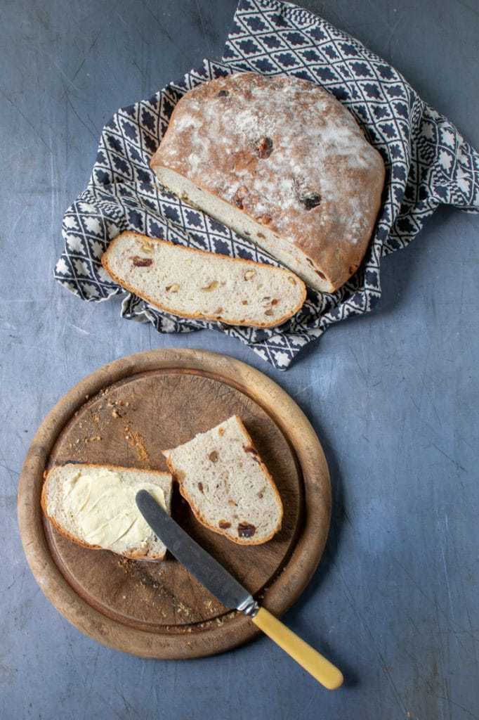 Overhead picture of a rustic date and walnut loaf of bread, with two slices on a bread board being spread with butter. Recipe. 