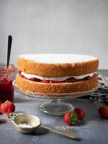 Vegan Victoria sponge cake on a cake stand with strawberry jam next to it. Get the recipe.