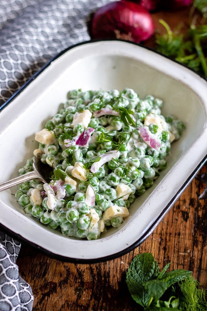 Creamy pea salad recipe served in an enamel dish with a grey napkin and fresh herbs scattered on the side, plus a red onion. 