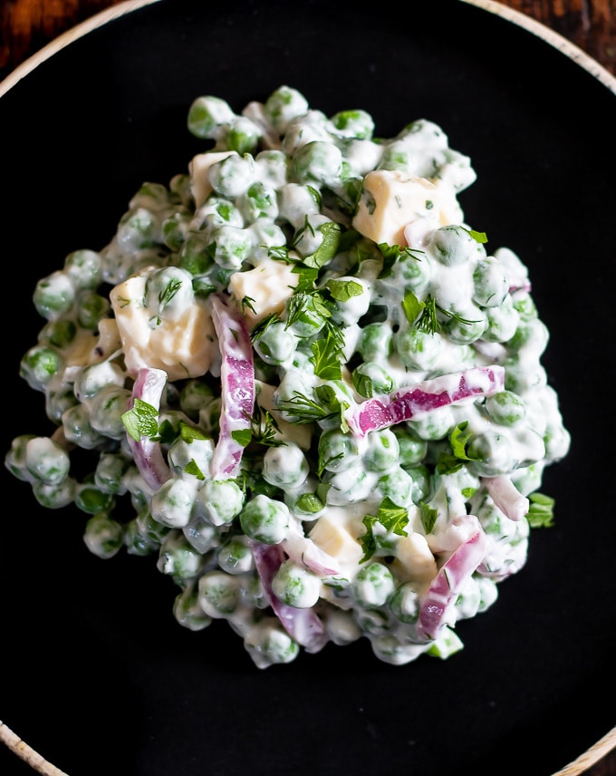 Close up of a scoop of creamy pea salad recipe, with red onion, cheese and fresh herbs on a black plate. 
