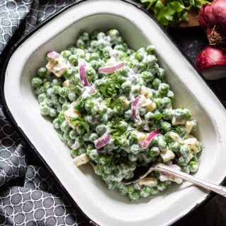 A bowl full of creamy pea salad. Next to black and white napkin with fresh herbs and red onion on a chopping board on the side.