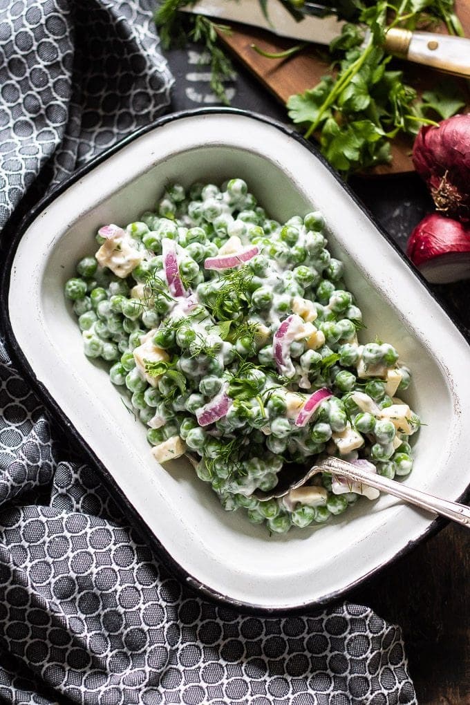 A bowl full of creamy pea salad. Next to black and white napkin with fresh herbs and red onion on a chopping board on the side. 