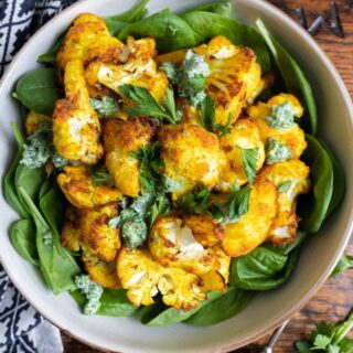 A bowl of bright yellow turmeric roasted cauliflower on a bed of spinach topped with a fresh herb dressing.